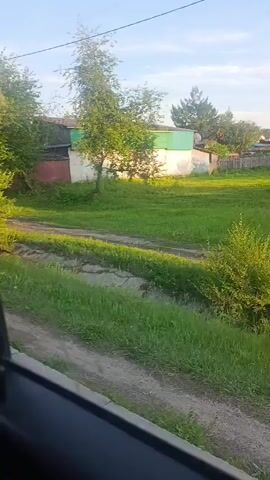 The video shows a view from a moving vehicle, likely a car, looking out a window. The scene is a rural or suburban area with green grass, trees, and some small buildings in the background. The sun is shining, casting shadows. The vehicle appears to be traveling on a dirt road or path. The video is shaky due to the movement of the vehicle.