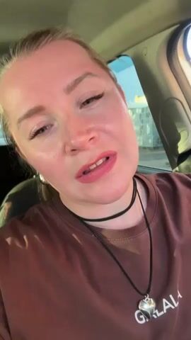 A woman with blonde hair pulled back is sitting in the driver's seat of a car, looking at the camera. She is wearing a brown t-shirt and a black choker necklace with a silver heart pendant. The woman is speaking, and her expression changes from a serious look to a smile. The sun is shining through the car window, casting a shadow on her face.