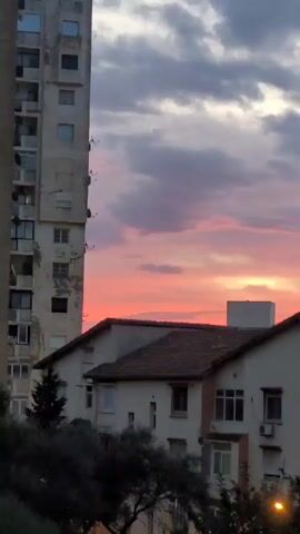 The video shows a sunset sky with pink and purple hues, partially obscured by gray clouds. The foreground includes a tall, weathered concrete building with numerous windows and balconies. To the right, there are two single-story houses with white walls and brown tiled roofs. A few trees with dark green foliage are visible at the bottom of the frame. The setting suggests an urban or suburban area at dusk.