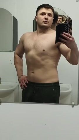 A shirtless man in green sweatpants stands in a bathroom, taking a selfie in a large mirror. The bathroom has white walls and several sinks with arched mirrors above them. A small, rectangular mirror is mounted to the left of the main mirror. The man is fair-skinned with short dark hair. He flexes his left arm and poses, holding his phone in his right hand. Several other men can be seen reflected in the mirror behind him.