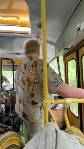 A person wearing a floral-patterned shirt and light-colored pants is standing inside a yellow vehicle, possibly a bus or tram, holding onto a yellow pole and handrail. The person's back is to the camera, and they appear to be looking out a window on the right side of the vehicle. Through the windows, lush green trees and foliage are visible, suggesting the vehicle is moving through a natural, possibly wooded, area. The interior of the vehicle is also yellow, with additional poles and seating visible. A bag is on the floor near the person. The video is shaky, indicating movement.