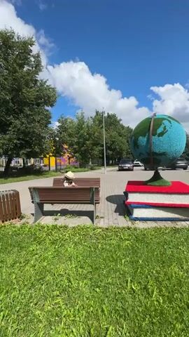 The video shows a person wearing a hat sitting on a park bench in front of a path and trees. There is a large globe decoration and stacked books made of flowers to the right of the bench. The person is facing away from the camera, with their head down. The sun is shining, and there are clouds in the blue sky. There are cars driving on a road in the background.