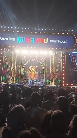 The video shows an outdoor concert with a large crowd facing a brightly lit stage. The stage has a large screen showing the performers. The sign above the stage reads "TURKIYE KULTUR YOLU FESTIVALI," with "TURIZM BAKANLIGI" displayed on an upper screen. The crowd is densely packed, and some individuals are holding up their hands and cheering. The lighting is dramatic, with spotlights and illuminated panels framing the stage. The overall environment suggests a festive atmosphere.