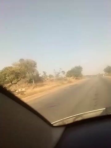 The video shows a view from inside a car driving on a straight asphalt road. The roadside vegetation consists of dry, scrubby plants and a few scattered trees. A motorcyclist wearing a yellow shirt and helmet drives towards the car on the same side of the road. The sky is a hazy blue. The interior of the car is visible in the foreground, showing the dashboard and a small section of the windshield. The overall atmosphere is dusty and somewhat arid.