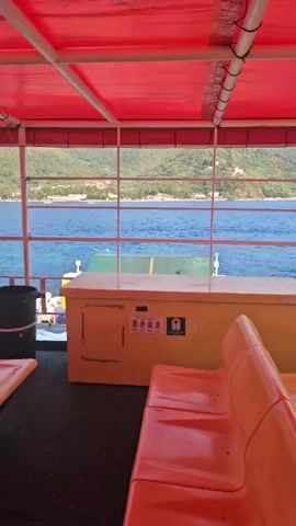 The video shows the interior of a ferry boat with orange seating. The boat is moving, as evidenced by the wake visible in the water. The ferry is passing by a coastline with buildings and trees. The sky is clear and the sun is shining. The video appears to be shot from the perspective of a passenger on the ferry.