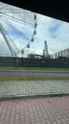The video depicts an amusement park, likely Energylandia. A large Ferris wheel with white carriages and the Energylandia logo is prominently displayed. Behind the fence and a strip of grass is the wooden construction of a roller coaster. The weather appears overcast. The camera is moving, possibly from a vehicle, as the scenery passes by. A small bus stop shelter is seen. A paved brick area is in the foreground.