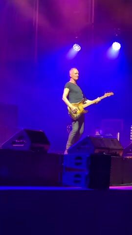 A man is playing a bass guitar on a stage. He is wearing a dark blue t-shirt and grey pants. He is bald and has a tan complexion. He is standing on a stage with purple and blue lighting. There are speakers on the stage. He is looking to his left.