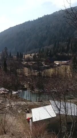The video shows a mountainous landscape in early spring or late winter. The foreground consists of bare trees and shrubs with some dried vegetation. A river or stream winds through the scene, and there are several buildings, likely houses, scattered among the trees. In the distance, a large mountain covered with trees dominates the background, with patches of blue sky visible above.  The scene is shot from a slightly elevated perspective, giving a broad view of the valley.