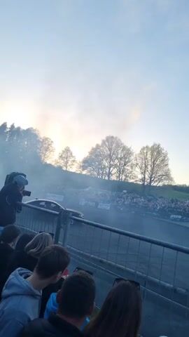 The video shows a black car drifting on a track surrounded by spectators. A metal fence separates the crowd from the track. The background consists of a grassy hill dotted with trees against a light blue sky tinged with the setting sun. Heavy smoke from the drifting car fills the air. The crowd includes people of varying ages and genders. Some are recording the event with their phones. The overall scene captures a dynamic moment at a drifting competition.