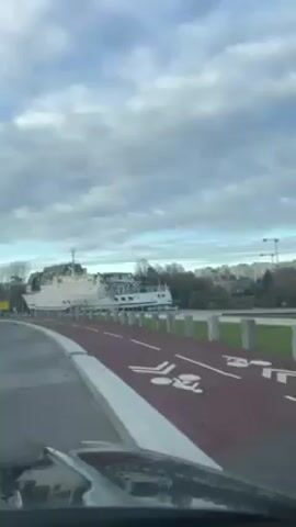 The view from inside a vehicle driving past a large white boat parked on a riverbank. A red bike lane is on the right, with white stone barriers separating it from the grassy area. Beyond the boat, there are apartment buildings and a construction crane in the distance. The sky is overcast with thick clouds. The camera pans to the right, showing more of the grassy area and some shipping containers.