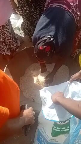 Here's a detailed description of the video:

Several people are gathered around a large metal bowl filled with rice or grain. One person, wearing a black headscarf and patterned clothing, is scooping the rice from the bowl into a white plastic bag using a white plastic container. The bag is labeled "THE HOME of GOOD FOOD." Other individuals are standing nearby, including one person in an orange shirt. The scene appears to be outdoors in sunlight. The ground is light-colored and dusty.