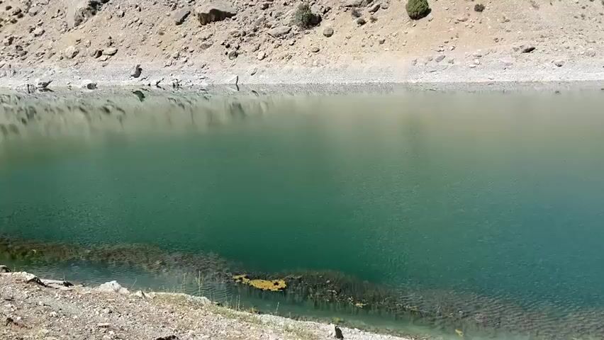The video captures a wide, still body of clear, turquoise water. The water is bordered on one side by a steep, rocky embankment composed of beige and grey stones, with sparse, dry vegetation. On the opposite side of the water, a rocky shore is visible with some green vegetation emerging from the water. The sun appears to be shining, casting a soft light on the scene and creating a gentle reflection on the water's surface. The overall impression is one of a serene, natural landscape.