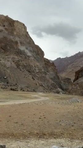 The video pans across a barren, mountainous landscape under an overcast sky. The foreground is a dusty, dry terrain with sparse vegetation and rocks. A winding dirt path is visible. To the left and right, steep, rocky mountains rise, casting shadows. The mountains are composed of layered rock formations in shades of brown and grey. In the distance, the valley narrows, with more rugged peaks visible. There are no people or visible animals in the scene.