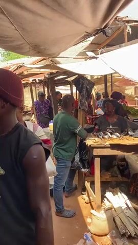 The video shows an outdoor market scene, possibly in Africa, with stalls made of wood and covered by tarps. Several people are visible, mostly women, dressed in colorful clothing. In the foreground, a person wearing a pink shirt with heart patterns is looking towards the camera. To their right, a woman is tending to a stall displaying several small fish on a wooden surface. Behind her, other people are interacting, and some are carrying goods. The ground appears to be dirt. The overall atmosphere suggests a busy marketplace.