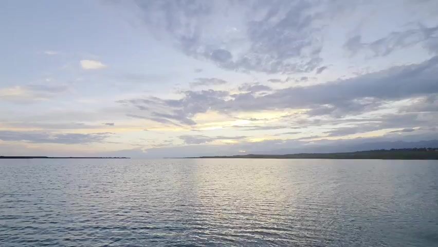 This video shows a wide shot of a large body of water, likely a sea or ocean, under a cloudy sky. The sun is setting, casting a soft glow on the water and clouds. The horizon is visible in the distance, with a faint silhouette of land. The water is calm with gentle ripples. The overall atmosphere is serene and peaceful.