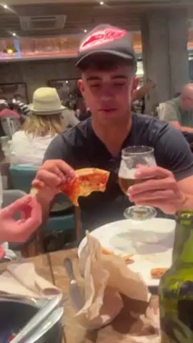 Here’s a forensic description of the video:

The video features a young man wearing a gray and red Detroit Red Wings baseball cap and a black t-shirt, sitting at a wooden table in a busy restaurant. He is holding a slice of pizza in his left hand, from which he has already taken a bite, and a glass of beer in his right hand. The table in front of him is cluttered with a white plate containing leftover pizza crusts, crumpled paper napkins, a butter knife, and a green beer bottle.  Other people can be seen in the background, including a woman wearing a wide-brimmed hat. The man is speaking and looking directly at the camera, alternating between appearing surprised and smiling, as he is prompted to say something to the camera.
