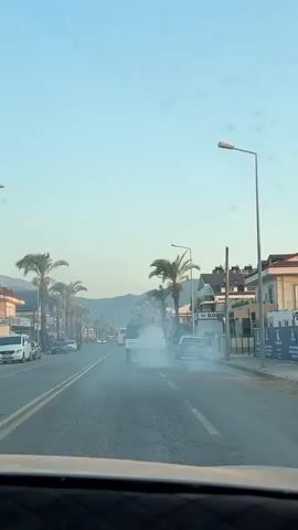This video captures a street scene in Fethiye, Turkey, likely during the day given the bright sky and lighting. The shot is taken from inside a vehicle, looking forward down a road lined with palm trees and buildings. The primary activity is a small white truck emitting a large cloud of white smoke or fog from its rear. The smoke billows out, obscuring the truck and a portion of the road behind it.

Other vehicles are visible on the road, including cars, a bus in the distance, and parked vehicles along the sides. The buildings lining the street appear to be shops or businesses, with one sign clearly displaying "KOO" followed by what might be other letters. The architecture suggests a Mediterranean or coastal resort town. The presence of palm trees and the clear sky contribute to a warm weather atmosphere. The smoke from the truck is a dominant visual element, creating a hazy effect and drawing attention to the truck's activity, which could be pest control, street cleaning, or some other fogging operation. The overall mood is one of routine street activity disrupted by the visible emission from the truck.