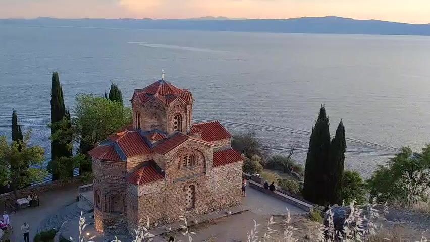 This aerial video captures the Church of St. John at Kaneo in Ohrid, North Macedonia, overlooking the vast, calm waters of Lake Ohrid. The time of day appears to be late afternoon or early evening, with soft, warm light illuminating the scene and casting gentle shadows. The sky is partly cloudy, with a gradient of soft pinks and oranges near the horizon, suggesting a sunset or sunrise.

The primary subject is the ancient stone church, perched dramatically on a cliff overlooking the lake. Its distinctive Byzantine architecture features a central dome, red-tiled roofs, and arched windows. The church is surrounded by lush green cypress trees, some of which are tall and slender, framing the structure.

In the foreground, at the base of the church, there are a few people visible. Some are seated at tables, possibly enjoying the view, while others are walking around the church grounds. Their presence suggests a tranquil atmosphere, with visitors enjoying the historical site and its breathtaking natural surroundings.

The lake itself is a prominent feature, its surface rippling gently, with a boat creating a noticeable wake in the distance. The far shore of the lake is lined with mountains, their peaks softened by the distance and the atmospheric light. The overall mood is one of peace, beauty, and historical reverence. There is no visible text in the video.