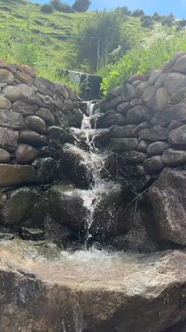 A small waterfall flows over rocks, surrounded by green foliage and a grassy hillside under a clear blue sky.