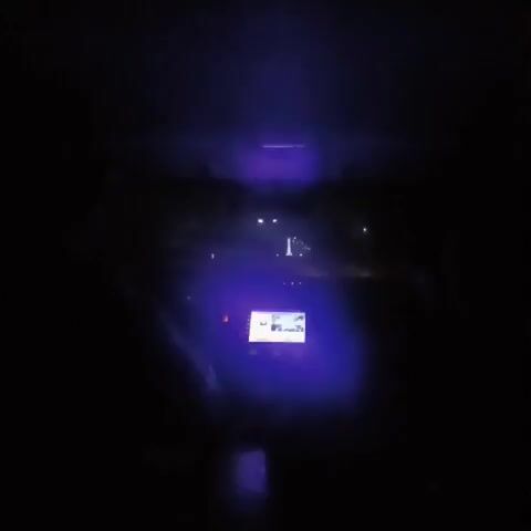The video shows the interior of a car at night. The dashboard is illuminated with a blue light, and the car's navigation screen is visible. Through the windshield, streetlights and buildings can be seen in the distance. A single windshield wiper can be seen moving from the driver's perspective.  The car appears to be moving along a road.