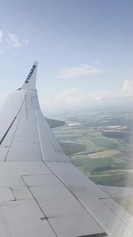This is a vertical video taken from the window of a Ryanair airplane in flight. The view is of the right wing of the plane, with the word RYANAIR clearly visible on the wingtip. Below the wing, the landscape is visible as a patchwork of green and brown fields, with some areas appearing to be cultivated. The sky is a clear blue with a few scattered white clouds. The camera appears to be slightly tilted, giving a sense of the airplane's movement.