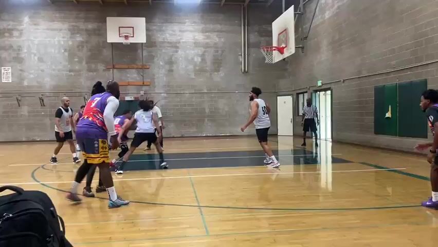 The video shows a basketball game in an indoor gymnasium. There are at least ten men on the court, some in white jerseys and some in purple and red jerseys. The court is made of polished wood with painted lines. There are two basketball hoops with backboards visible. One player in a purple jersey with the number 7 is dribbling the ball. Another player in a white jersey is defending him. A third player in a purple jersey is attempting a shot. The men appear to be of various ethnicities and ages. The walls of the gymnasium are made of concrete blocks, and there are some windows and doors visible. The lighting is artificial, coming from overhead fixtures. The scene is dynamic, with players moving and interacting during the game.