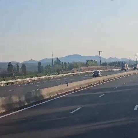 The video shows a convoy of emergency vehicles, primarily fire trucks, driving on a highway. The vehicles are moving in the same direction. The highway has multiple lanes and a concrete barrier separating the lanes. In the background, there are trees, hills, and a clear sky. The sun appears to be shining, suggesting it is daytime. The vehicles are equipped with flashing blue lights, indicating they are responding to an emergency. The overall scene suggests an emergency response operation taking place on a highway.
