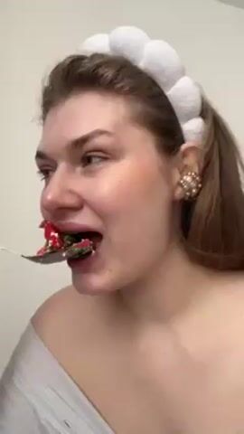 A woman with light skin, brown hair, and a white puffy headband is eating food from a spoon. She chews the food, closes her eyes in what appears to be enjoyment, and slightly puckers her lips. She is wearing a light grey off-the-shoulder top and large, decorative earrings. The background is a plain white wall. The audio includes dance music.