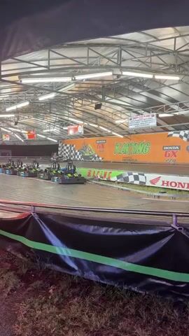 The video shows an indoor go-kart track. The track is lit by overhead fluorescent lights. The walls of the track are decorated with orange and green banners that say "Karting" and "Honda GX 270". There are also checkered flags and logos of sponsors. Several go-karts are visible on the track, some of which are in motion. The go-karts are low to the ground and appear to be designed for racing. The track has banked turns and straightaways. The video appears to be taken from a slightly elevated position, possibly from a spectator area. The overall atmosphere is one of an indoor amusement park or racing facility.