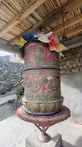 A large, ornate Tibetan prayer wheel is shown in slow rotation. The wheel is cylindrical and decorated with intricate carvings and red and gold panels. Colorful prayer flags are draped over the top. The wheel is housed under a rustic wooden roof supported by beams. The background shows stone walls and a dirt ground. The video is shot from a stationary, medium-distance perspective.