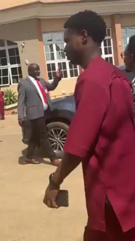 The video shows a group of people outdoors near a building and a car. Several individuals are walking, including a man in a suit and red tie walking near a gray SUV. A woman with a red purse stands next to him. Other people are walking in the same direction, including two younger men. One is wearing a maroon shirt, and the other is wearing a black and white patterned jacket. The building behind them has a light-colored facade and multiple windows. The area appears to be paved.