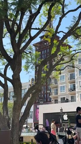 A video shows a tree with green leaves in the foreground. Behind the tree, a tall building with a clock tower can be seen. To the right of the building, there is another building with balconies and windows. In front of this building, there is an Apple Store with a large white apple logo. Several people are visible in the foreground, including a woman with a stroller and a child. The overall environment appears to be an outdoor shopping area on a sunny day.
