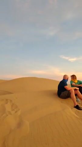 A group of at least nine people is sitting on a sand dune, possibly in a desert. A red dune buggy is parked nearby. In the distance, more sand dunes are visible. The sky is filled with clouds and the colors of a sunset.  Some people are talking to each other.  Others are looking out at the view. The scene appears peaceful and relaxed.