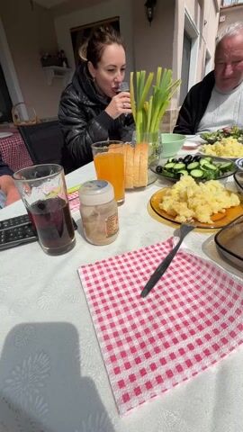 The video depicts a family enjoying a meal outdoors on a sunny day. The camera focuses on a table laden with food, including potatoes, salad, hard-boiled eggs, and bread. Drinks like juice and dark soda are visible. People are seen eating and drinking around the table. The camera then pans to show a garden area with a gazebo and a television. The background reveals a well-maintained lawn, other buildings, and a view of hills in the distance.