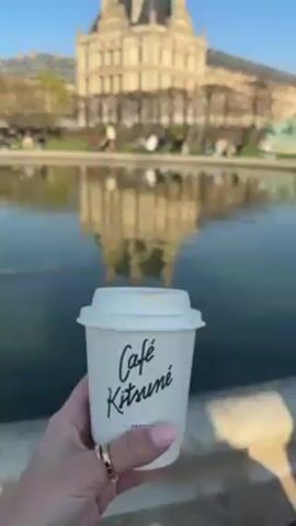 The video shows a person holding a Café Kitsuné paper cup in front of a body of water reflecting a large building. The building is likely a historical landmark, possibly a palace or museum. People are sitting along the edge of the water, enjoying the scenery. The sky is clear and blue.