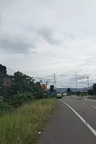 The video captures a wide shot of a road in Payakeureuleh, Indonesia, under an overcast sky. The time of day appears to be daytime, with soft, diffused light due to the dense grey clouds.

On the left side of the frame, there is a grassy area leading to a sidewalk. A prominent yellow diamond-shaped road sign with a black pedestrian crossing symbol is visible. Further along the left, there are trees, bushes, and a smaller blue rectangular sign whose text is illegible. Multiple utility poles and power lines stretch across the sky, with a large, imposing power transmission tower (pylon) dominating the mid-ground.

The road itself is a multi-lane asphalt surface with clear white lane markings. Several modern streetlights with double arms are positioned along the right side of the road. In the distance, a yellow truck is seen slowly driving away from the camera on the right-hand lane. The right side of the road also features a median or road island marked with black and white bollards, and more green foliage.

In the far background, hazy mountains or hills are visible, blending into the cloudy horizon. The overall scene depicts a functional road infrastructure in a natural, somewhat rural setting. No specific human activity beyond the truck's movement is clearly visible.
