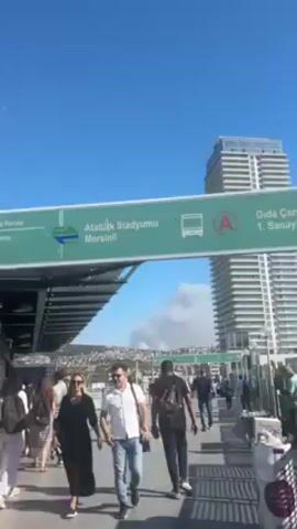 Here is a detailed description of the video:

The video captures a view from an elevated walkway or bridge, likely part of a transportation hub, based on the signs. The main focus is a large plume of smoke rising in the distance against a clear blue sky. The smoke is billowing, suggesting a significant fire or industrial incident.

In the foreground, a green sign provides information about a transit stop. It mentions "Atatürk Stadyumu" and "Mersinli," along with a symbol for a bus stop and what appears to be a route designation "A." 

A crowd of pedestrians is walking along the walkway. The people appear to be dressed in casual attire. A tall, modern skyscraper is visible to the right of the walkway.

The video appears to be taken on a bright, sunny day, with clear visibility.