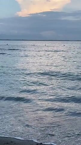 The camera is stationary at a beach, capturing a calm sea with small waves gently lapping at the shoreline. The sky is overcast, with hints of pale orange and pink where the sun might be setting or rising. In the distance, there are several buoys visible on the water. The beach itself appears sandy, with small pebbles visible near the waterline.