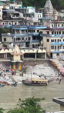 The video presents an elevated, distanced view of a section of shoreline along a river lined with buildings and people. A body of water occupies the foreground, with several boats visible: one packed with passengers, and others docked along the sandy beach. On the beach, numerous individuals are present, some walking along the shore, others standing in small groups, and some swimming in the river. Adjacent to the shoreline, a tiered structure with white arched supports and a small temple-like building on top can be observed. Behind this structure, a dense arrangement of buildings stretches upwards, with varying levels, colors, and architectural styles. The buildings are tightly packed together, creating an urban landscape that rises along the riverbank. Some buildings have signs in what appears to be Hindi script. The overall scene depicts a lively riverside environment with a mix of recreational activities and urban density.