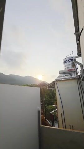 The video captures a serene view of either a sunrise or sunset over a mountainous landscape, filmed from an elevated vantage point, possibly a balcony or rooftop.

The initial shot reveals a panoramic scene: in the distance, lush green mountains are silhouetted against a bright, hazy sky, with the sun peeking just above their peaks, casting a soft, warm light. To the right, a part of a building is visible, featuring a large water tank on its roof. The tank is branded with "TÂN" and "ARISTON" and is connected to what appears to be a solar water heating system, with various pipes running along the building's exterior. A plain white wall of another structure occupies the left side of the frame.

The camera then smoothly zooms in on the sun and the mountains. The sun appears as a distinct, bright disc, partially obscured by the mountain ridge and surrounded by a soft, diffused light. Green foliage is visible in the mid-ground below the mountain.

The final segment offers a close-up of the sun, which appears as a brilliant, almost white orb against a yellowish, hazy sky, with subtle cloud formations around it. Only a small portion of the mountain's silhouette remains visible at the very bottom of the frame.

No people are present in the video. The lighting suggests either early morning or late afternoon, with a generally hazy but bright atmosphere. The objects include residential fixtures like a water tank and solar heater, set against a backdrop of natural mountainous scenery, aligning with the Bac Natural Heritage Area Trang, Vietnam context.