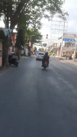 The video captures a street scene, possibly in a developing country, with a focus on vehicular traffic and surrounding buildings. The viewpoint appears to be from a moving vehicle, likely a motorcycle, judging by the low angle and the presence of a motorcycle in front and to the left.

In the foreground, the asphalt road stretches ahead. A black motorcycle with a rider wearing a dark jacket and helmet is moving away from the camera in the middle of the lane. Further ahead, a white car is also moving in the same direction.

To the left of the road, there are several structures. A large tree with leafy branches dominates the upper left portion of the frame. Below the tree, a building with a dark facade and an awning is visible, with a motorcycle parked in front of it. Further down the street, there are more buildings with shops and signs, some of which are partially obscured by trees and poles. One sign appears to have Indonesian text. Colorful flags or banners are hanging from poles.

To the right of the road, there are more buildings, including a store with a prominent blue and white sign that reads "Indomaret," a common convenience store chain in Indonesia. The buildings on this side appear to be more modern, with multiple stories and balconies. A large, tall billboard structure, seemingly empty, stands against the sky.

Traffic lights are visible at an intersection ahead. At the beginning of the clip, the lights appear to be yellow, and then they change to red. This indicates that the traffic is about to stop. Overhead power lines crisscross the sky, strung between poles along the street. The sky is overcast with some patches of blue. The overall impression is a bustling street scene during daylight hours.