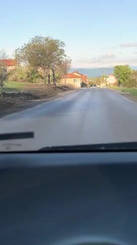 The view from inside a car traveling on a quiet, paved road through a rural area. The road is straight and has buildings and trees on either side. The sky is blue with some light clouds. The foreground shows the dashboard and the bottom edge of the windshield.  The road surface appears slightly wet or reflective.