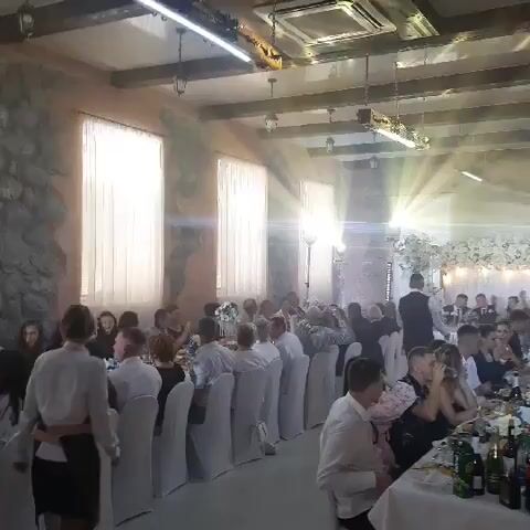 The video shows a wedding reception or a similar celebratory event taking place indoors. Long tables are set up, draped with white tablecloths and adorned with floral arrangements and bottles of drinks. Many guests are seated at the tables, engaged in conversation, eating, and drinking. The room is decorated with white floral arrangements and white fabric draping along the back wall, creating a festive atmosphere. Lighting beams are visible, suggesting spotlights or stage lighting, and the ceiling has exposed wooden beams and industrial-style light fixtures. People are dressed in formal or semi-formal attire. The overall impression is one of a lively and happy gathering.