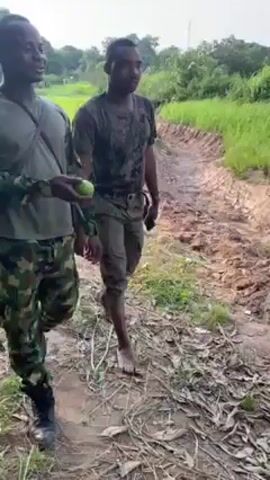 Two men are walking alongside a muddy ditch or trench. One man wears camouflage pants, a long-sleeved camouflage shirt, and black boots, and holds a green fruit-like object in his hand. The second man wears dark-colored pants and a dark shirt that appears wet and stained with mud, and is barefoot. He carries a black cell phone. The surrounding environment consists of tall green grass, trees, and dense foliage.