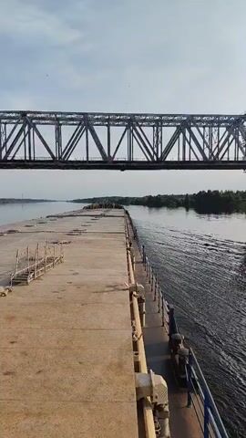 A vertical video captures a view from a vessel moving along a waterway towards a large metal railroad bridge spanning the water. The vessel's deck is visible in the foreground, with a concrete walkway extending to the left and a railing to the right. The water is rippling, indicating movement. Trees and vegetation line the banks of the waterway on either side, receding into the distance. The sky is clear with a few wispy clouds.