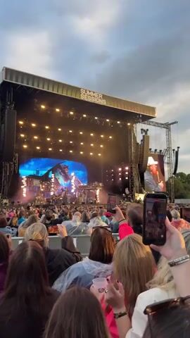 The video shows a crowd of people at an outdoor concert during the day. The stage is set up with a large screen in the background displaying a graphic of a rustic building with a blue sky. There are many lights on the stage and scaffolding. A band is performing on stage. The audience is seated in rows and many people are holding up their phones to record the performance. The sky is cloudy. The text "SUMMER SESSIONS" is visible on the top of the stage structure.