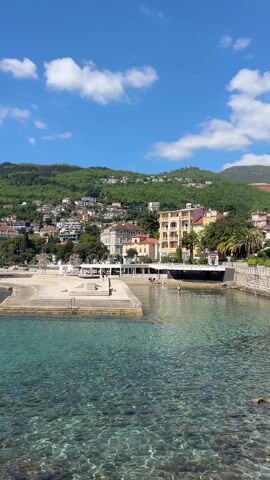 The video shows a coastal town in Croatia, with views of the waterfront, beaches, and nearby mountains. The water is clear and blue. People can be seen walking along the beach and swimming in the water. A bronze statue of a woman stands on a rock along the shore, overlooking the Adriatic Sea. The sky is blue with scattered clouds, indicating a sunny day. Buildings and villas are situated on the hillside, offering a scenic backdrop.
