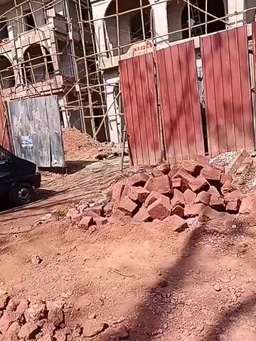 The video shows a construction site in an urban area. Two unfinished buildings with arched openings and scaffolding are visible behind a red corrugated metal fence. A pile of red bricks sits in front of the fence. A gray scooter is parked to the right of the fence, covered by a green tarp. A black car is parked on the left side of the frame. The ground is mostly dirt. The sky is blue and clear.