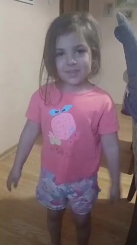 A young girl stands indoors in a room with light-colored walls and a light-colored wooden floor. She is wearing a pink T-shirt with a cartoon fruit design, and colorful patterned shorts. The girl looks directly at the camera and is smiling. There is a piece of furniture, possibly a table or desk, in the background to her right. The furniture is covered with a blue blanket or cloth, and a stuffed animal or other soft object rests on top of it. The girl momentarily spreads her arms out and then brings them down in front of her. The audio includes music and the sound of a young voice speaking.