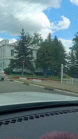 This is a dashcam video taken from a car driving through Zadonsk, Russia. The scene is a suburban street with trees, buildings, and other cars. The weather is sunny with some clouds, and the time of day appears to be afternoon. 

In the first frame, the car is parked, and the view shows a white gazebo with ornate railings and columns. The gazebo is located in a park-like setting with trees in the background.

As the video progresses, the car starts to move, and the view changes to the street. Several cars are visible, including a silver sedan and a gray SUV. The street is lined with buildings, some of which appear to be residential or commercial. There are also power lines and traffic lights visible. 

The video captures everyday life in Zadonsk, showing the flow of traffic and the typical scenery of a Russian town. No specific event is taking place, but the footage provides a glimpse into the local environment. The visible text includes license plates on some of the cars.
